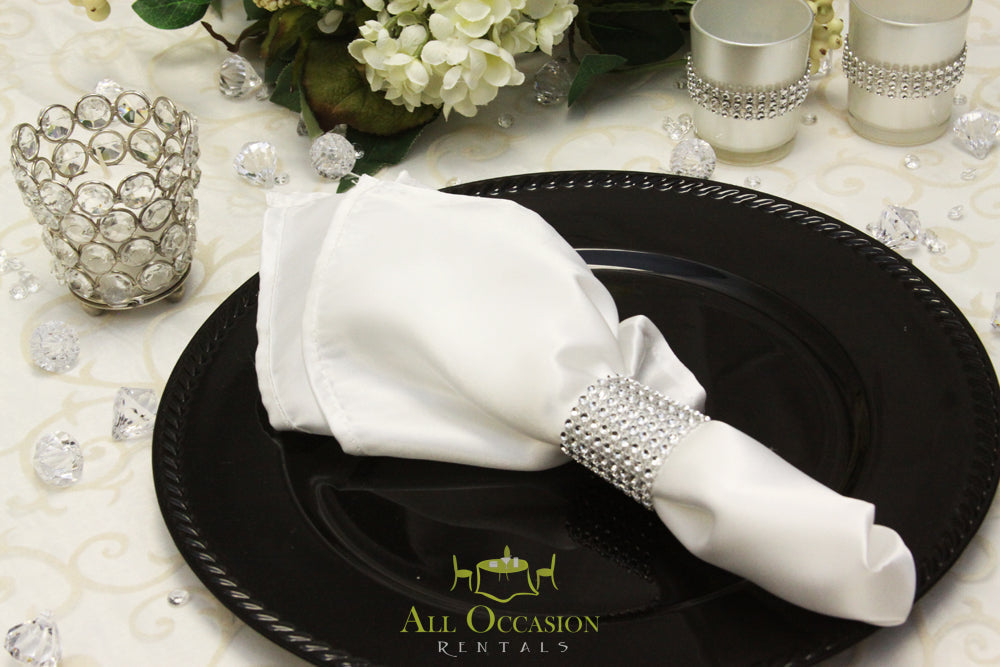 Elegant white satin napkin with jeweled ring on black plate; perfect for Corona event rentals and weddings.