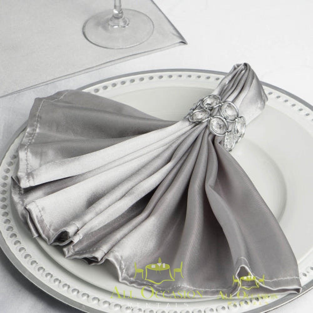 Elegant silver satin napkin with decorative ring on a white plate, perfect for Corona event rentals and weddings.